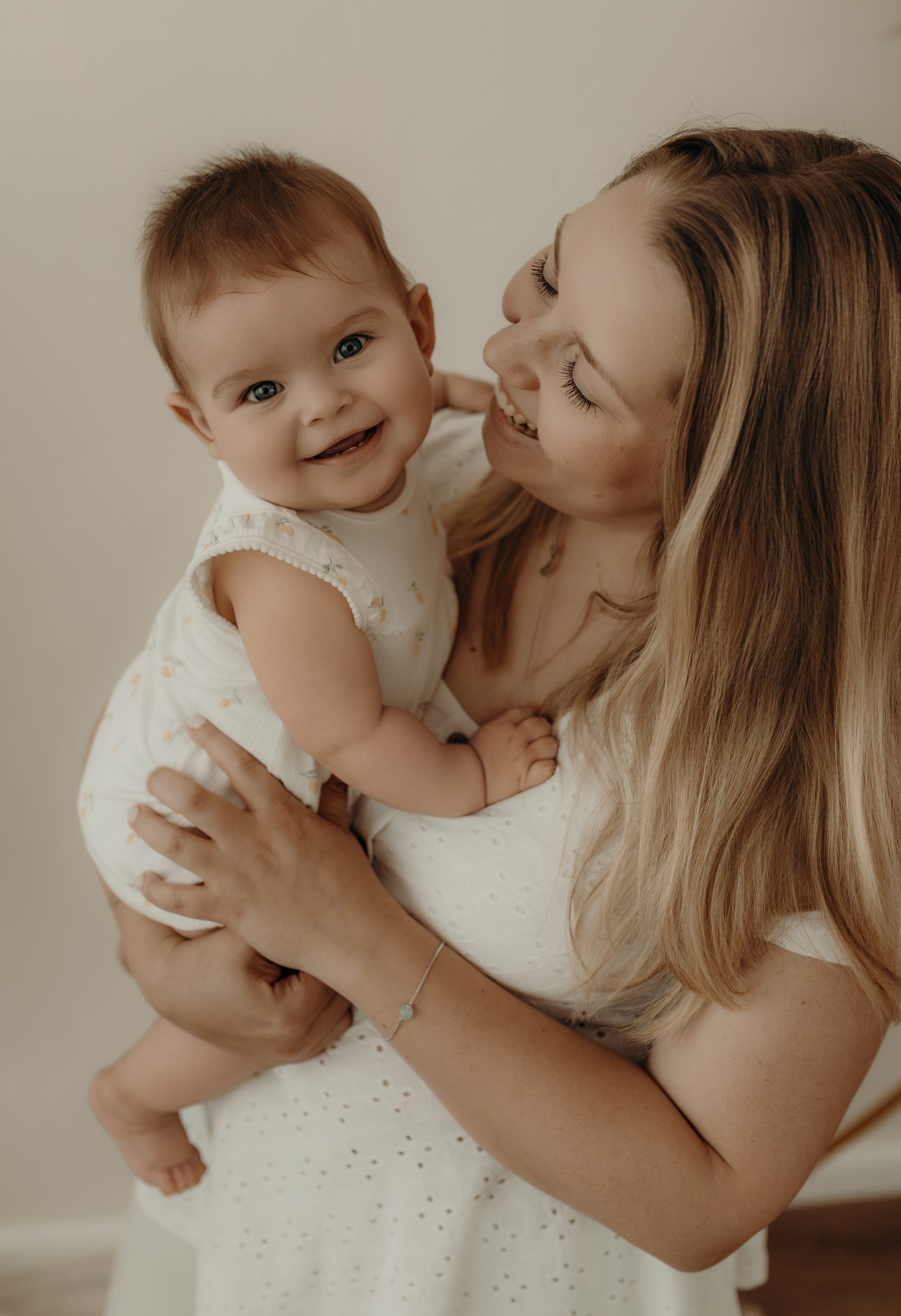 Mama hat Tochter auf dem Arm und lächelt sie an, das Mädchen lächelt in die Kamera