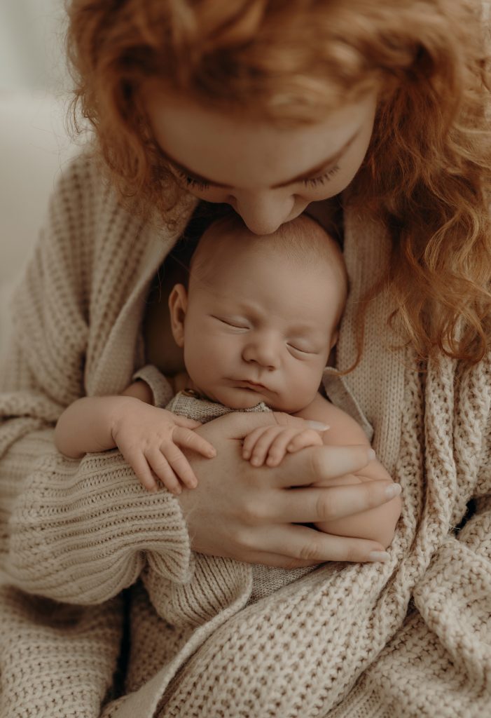 Baby ist im Arm der Mama in eine Strickjacke gekuschelt und die Mama küsst das Baby auf den Kopf